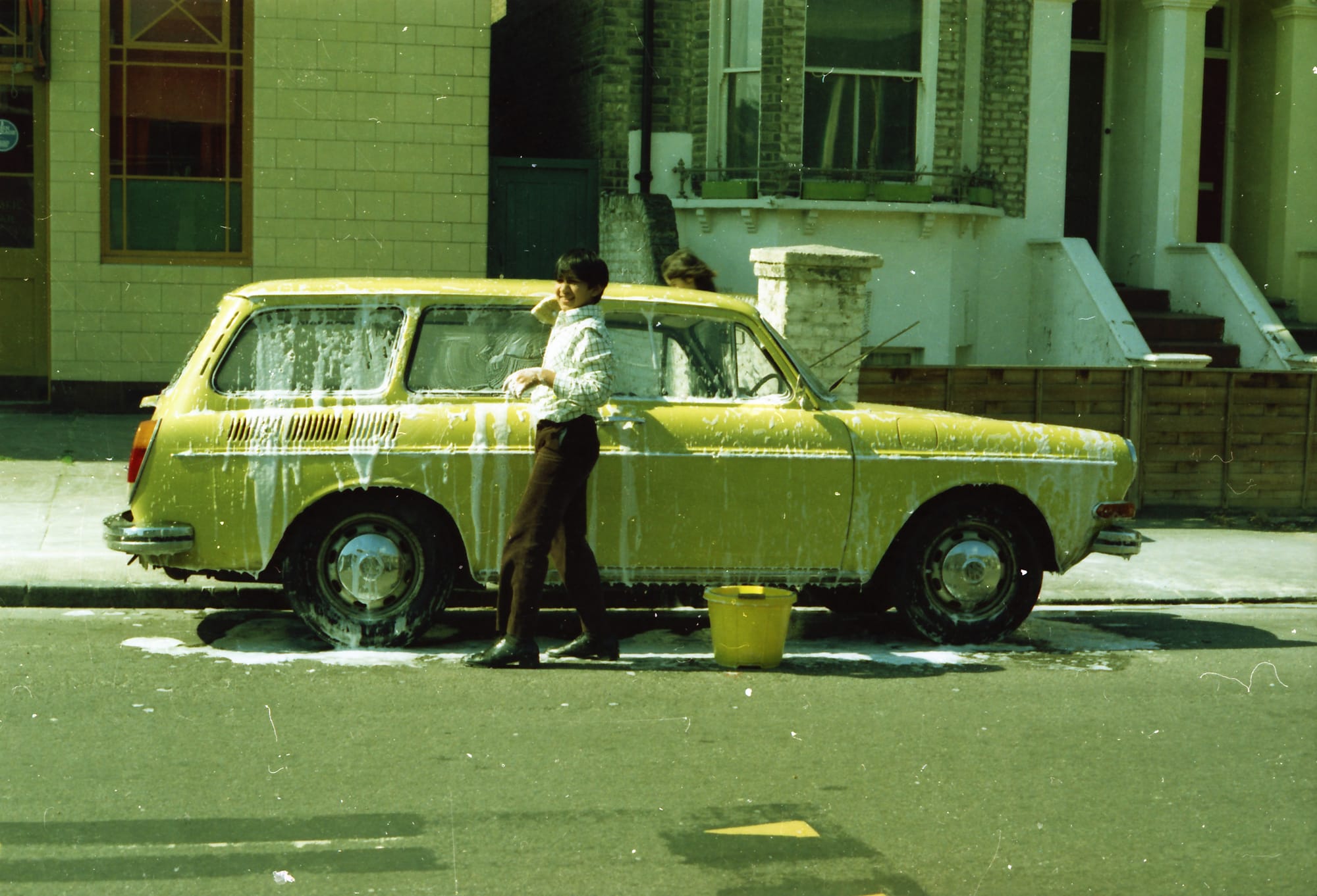 Ravi, leaning on the family's yellow VW Type 3 Variant, holding a sponge. A street in South London