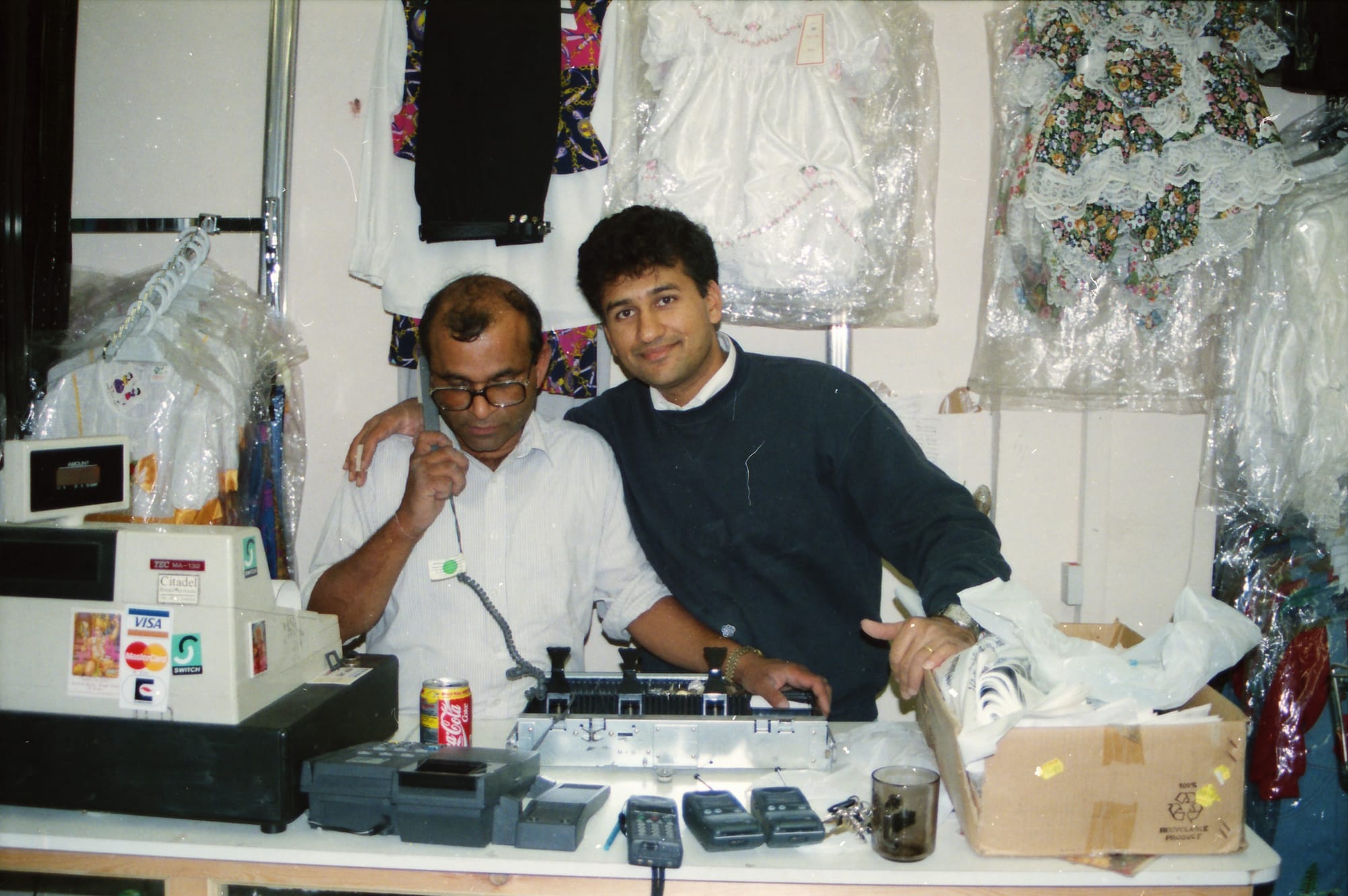 Sunil and a man on the telephone, behind the counter of a children's and babywear shop. A shop in Slough
