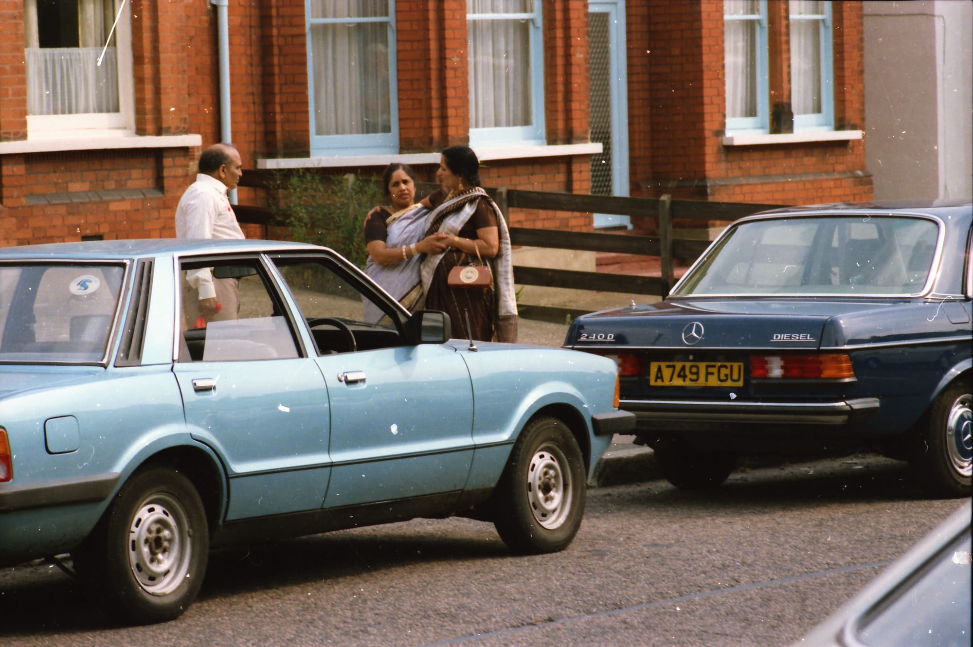 Two women in saris and a man. A terraced street in England