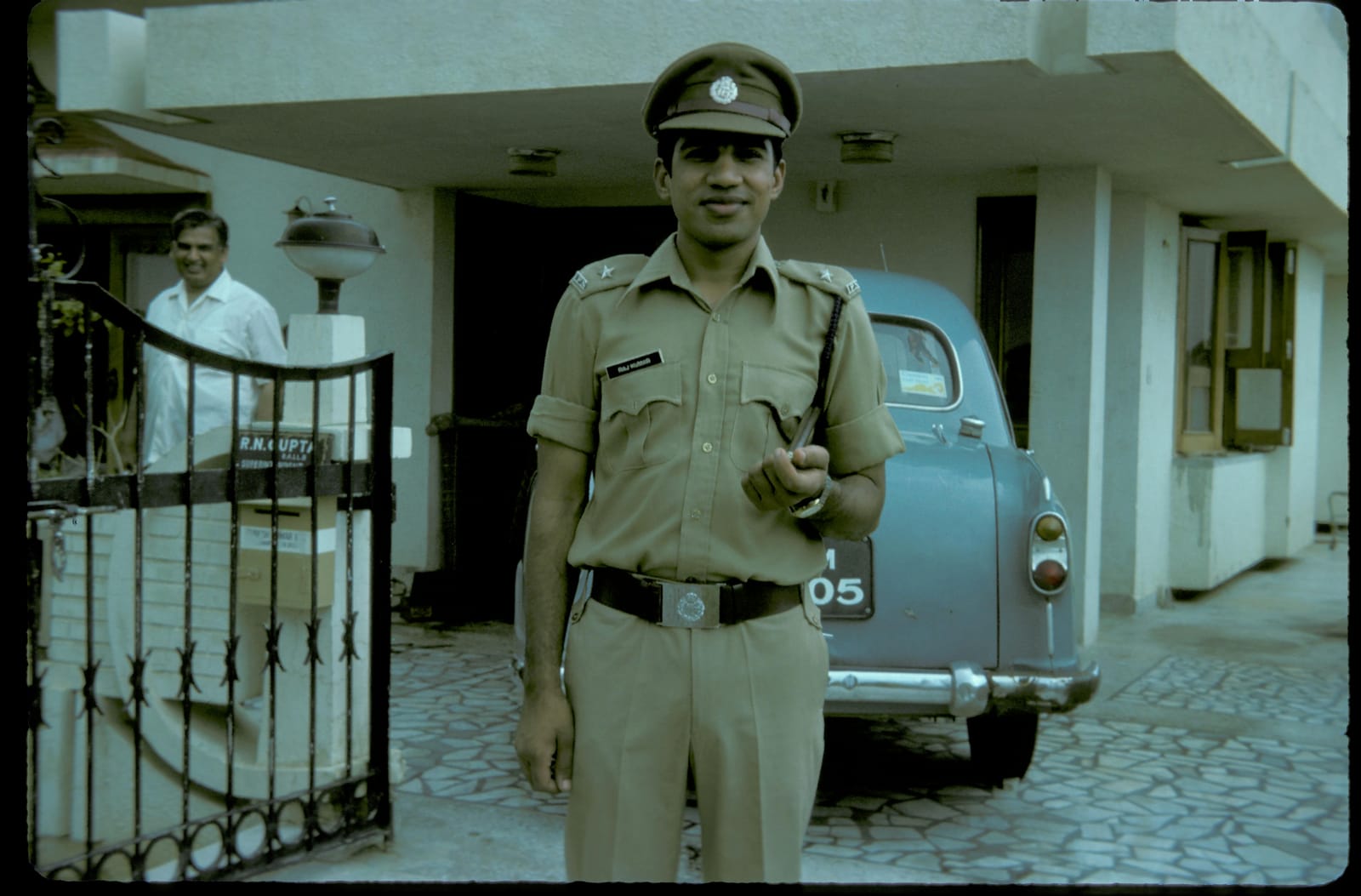Raj Kumar in full police uniform with cap and cane at a family gate, an Ambassador behind